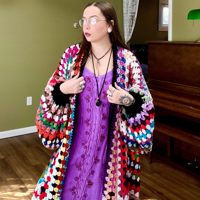 May include: A woman wearing a colorful crochet cardigan with a purple dress underneath. The cardigan is made of granny squares in various colors, including red, green, blue, and yellow. The woman is wearing a necklace with a pendant.
