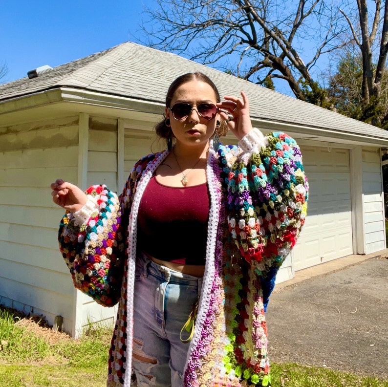 May include: A multi-colored crochet cardigan with a white trim and large, puffy sleeves. The cardigan is worn over a maroon tank top and blue jeans.