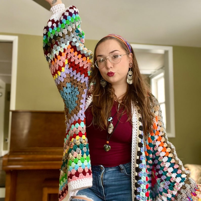 May include: A colorful crochet cardigan with a white border. The cardigan is made of granny squares in various colors, including red, blue, green, yellow, and brown. The person wearing the cardigan is also wearing a red shirt, blue jeans, and a headband.