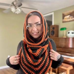 May include: A handmade crochet hood and scarf set in a striking orange and black striped pattern. The hood is designed to be worn over the head and shoulders, creating a cozy and stylish accessory. The scarf is triangular and complements the hood's design.