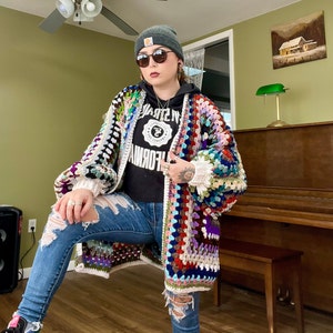 May include: A woman wearing a colorful crochet cardigan with a white crocheted hat and blue jeans. The cardigan has a variety of colors, including red, blue, green, and purple. The woman is standing in front of a piano.