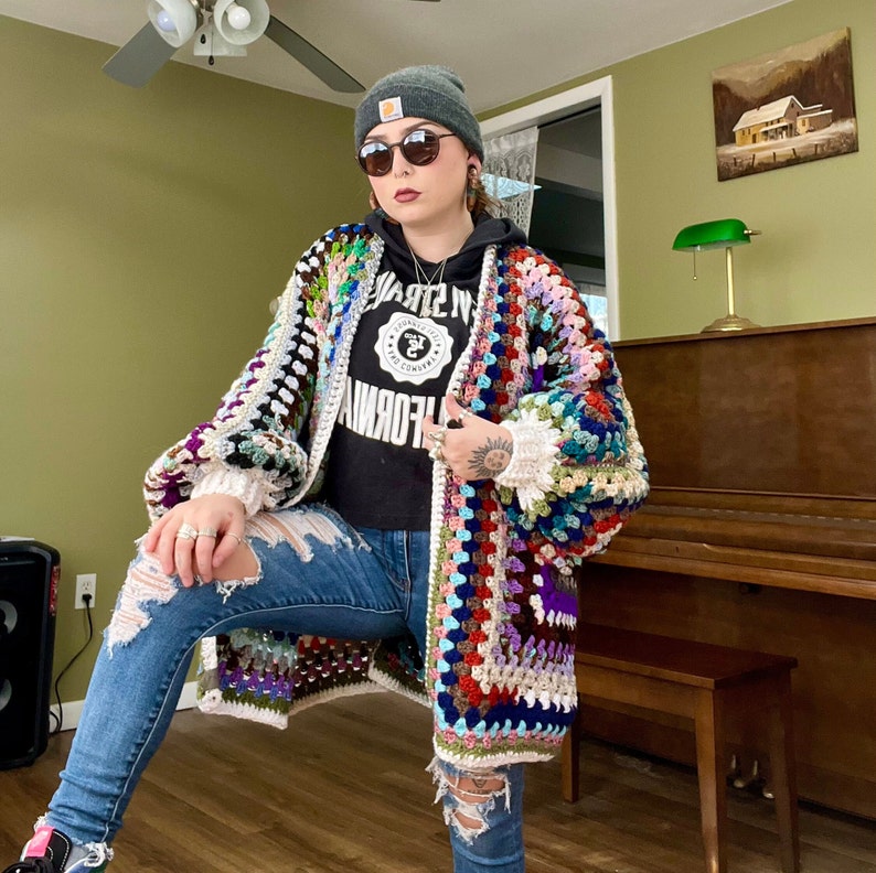 May include: A woman wearing a colorful crochet cardigan with a white crocheted hat and blue jeans. The cardigan has a variety of colors, including red, blue, green, and purple. The woman is standing in front of a piano.