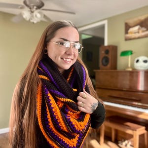 May include: A handmade crochet cowl scarf in black, orange, and purple. The scarf is triangular in shape and features a striped pattern. The yarn appears to be thick and soft, perfect for cooler weather. The cowl is worn around the neck and shoulders.