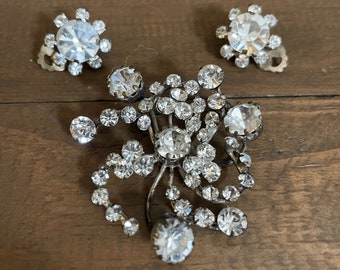 Può includere: Un set di gioielli vintage con una spilla e orecchini abbinati. La spilla ha un design floreale con molteplici strass chiari e scintillanti. Gli orecchini sono rotondi con un grande strass centrale circondato da pietre più piccole.