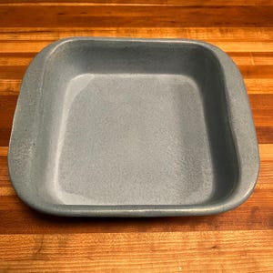 May include: A square, light blue ceramic baking dish with rounded corners. The dish has a speckled texture and is set on a wooden surface with a striped pattern.