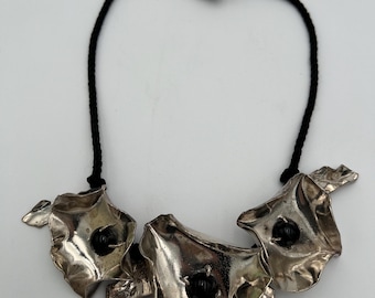 Puede incluir: Collar plateado con tres colgantes de flores estilizadas, con centros negros y detalles de hojas. Los colgantes están ensartados en un cordón negro, creando un diseño audaz y artístico. Una pieza de joyería única.
