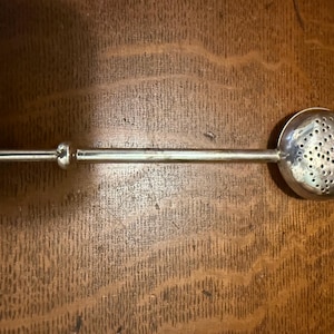 May include: A silver tea infuser with a perforated spherical basket and a long handle. The handle has a small ball at the end and a decorative ring near the basket. The infuser is designed for steeping loose leaf tea.