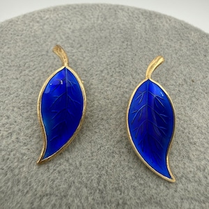 Puede incluir: Un par de pendientes con forma de hoja. Cada pendiente presenta un diseño de hoja de esmalte azul vibrante con ribete dorado. Las hojas tienen delicados detalles de veteado. Los pendientes están sobre un fondo gris.