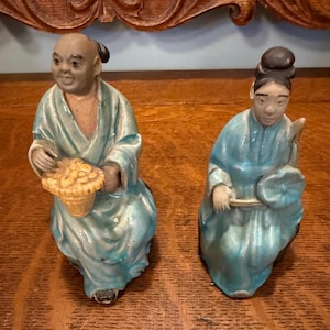 May include: Two antique ceramic figurines. One figure is holding a basket of fruit, the other is holding a fan. Both figures are wearing blue robes. The figurines are on a wooden surface.