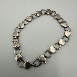 May include: A silver-toned metal bracelet composed of a series of heart-shaped links connected by small circular jump rings. The bracelet features a lobster claw clasp mechanism that is also shaped like a heart, securing the chain together.