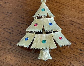 May include: Gold-toned Christmas tree brooch with red, green, and blue jewel accents. The tree is topped with a red star and has a textured surface. A festive accessory for the holiday season.