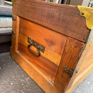 Puede incluir: Un ba&uacute;l de madera con protectores de esquina y herrajes de lat&oacute;n. El ba&uacute;l tiene forma rectangular con un asa en la parte delantera. La madera es de color marr&oacute;n rojizo.