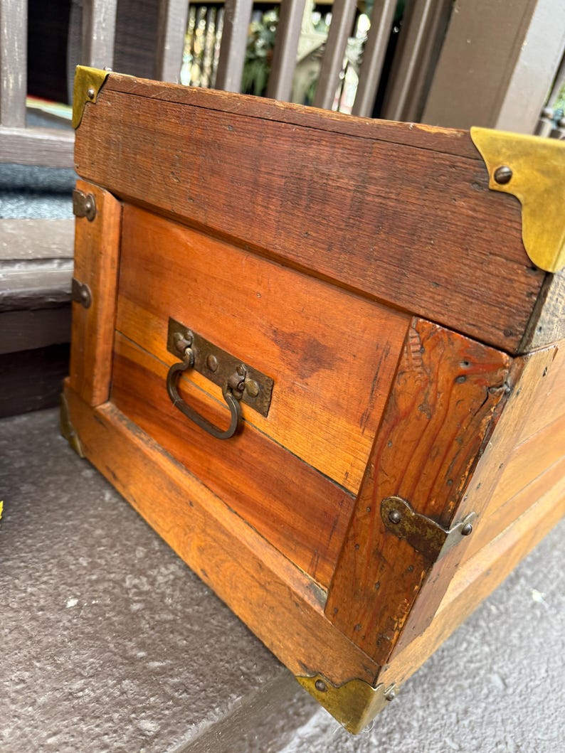 Puede incluir: Un ba&uacute;l de madera con protectores de esquina y herrajes de lat&oacute;n. El ba&uacute;l tiene forma rectangular con un asa en la parte delantera. La madera es de color marr&oacute;n rojizo.