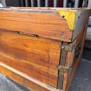 Puede incluir: Un ba&uacute;l de madera vintage con protectores de esquina y bisagras de lat&oacute;n. El ba&uacute;l es de madera marr&oacute;n claro con vetas visibles y forma rectangular. Los herrajes de lat&oacute;n a&ntilde;aden un toque de encanto antiguo.