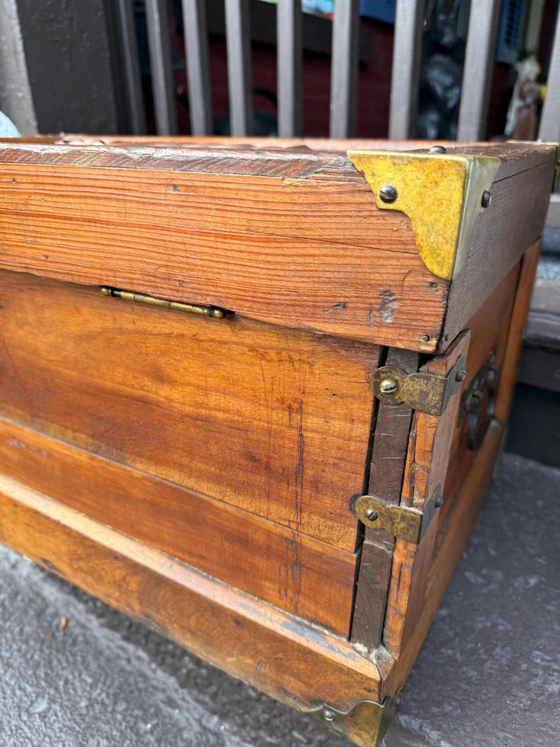 Puede incluir: Un ba&uacute;l de madera vintage con protectores de esquina y bisagras de lat&oacute;n. El ba&uacute;l es de madera marr&oacute;n claro con vetas visibles y forma rectangular. Los herrajes de lat&oacute;n a&ntilde;aden un toque de encanto antiguo.