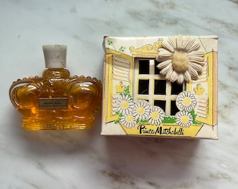 May include: A vintage perfume bottle shaped like a crown, with a label that reads "Crown Jewel". Next to it is a box with a window design and the words "Prince Matchabelli". The box is decorated with daisies.