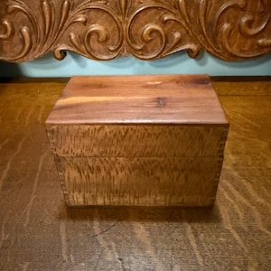 May include: A rectangular wooden box with a smooth, flat lid and a textured, tiger-striped grain pattern on the front panel. The box features visible finger joints at the corners, showcasing a natural light brown finish with darker wood grain details.