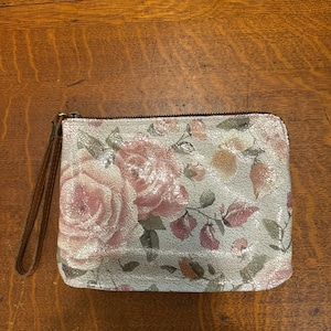 May include: A rectangular clutch with a floral pattern featuring pink roses and green leaves on a light gray background. The clutch has a brown wrist strap and a zipper closure.