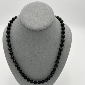 May include: A black beaded necklace displayed on a gray jewelry bust. The beads are faceted, reflecting light, and the necklace is a classic, elegant accessory. The background is a plain white.