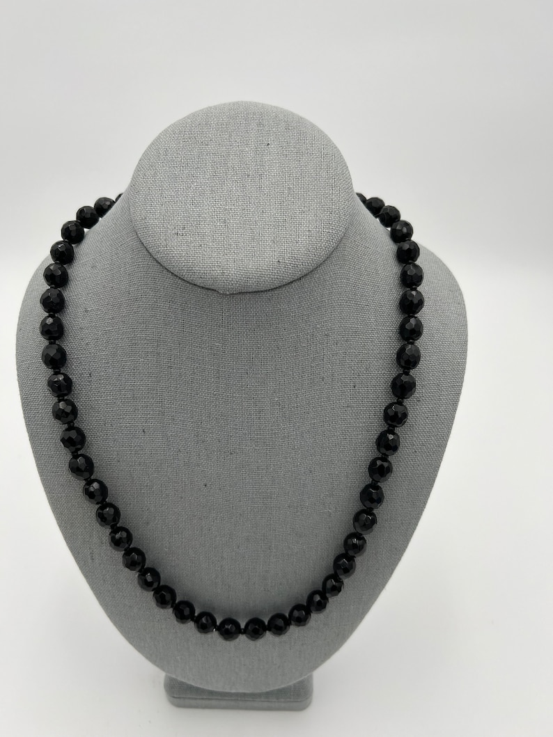 May include: A black beaded necklace displayed on a gray jewelry bust. The beads are faceted, reflecting light, and the necklace is a classic, elegant accessory. The background is a plain white.