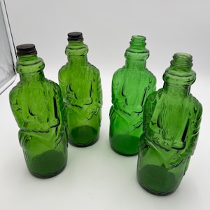May include: Four green glass bottles with a unique design. Each bottle has a detailed, embossed figure and a distinct shape. Two bottles have brown metal caps, while the other two have green caps. The bottles are arranged against a white background.