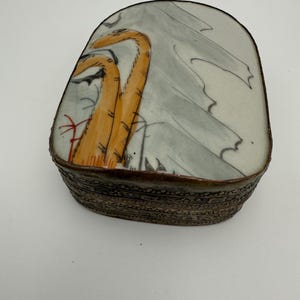May include: A ceramic trinket box with a lid featuring a hand-painted design. The lid has a light gray background with orange and black tiger-like figures. The box has a dark brown, textured base.