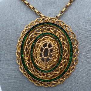May include: Gold-tone necklace with a large, circular pendant. The pendant features concentric circles of green stones and intricate gold rope detailing. The necklace chain is also gold-toned.