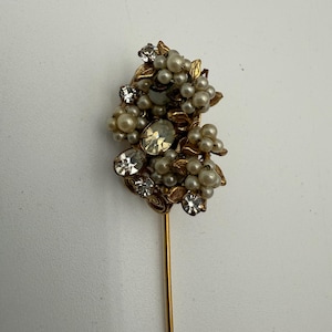 Puede incluir: Un broche de sombrero vintage con un racimo de perlas de imitación, pedrería transparente y hojas de metal dorado. El broche tiene un eje largo y delgado de color dorado. El diseño es ornamentado y elegante.