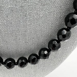 May include: A close-up of a black beaded necklace. The beads are large, faceted, and have a glossy finish. The necklace is displayed on a gray surface, highlighting the jewelry's dark color and texture.