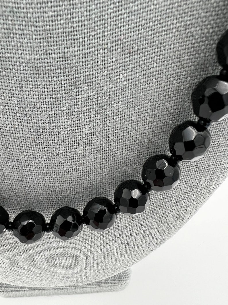 May include: A close-up of a black beaded necklace. The beads are large, faceted, and have a glossy finish. The necklace is displayed on a gray surface, highlighting the jewelry's dark color and texture.