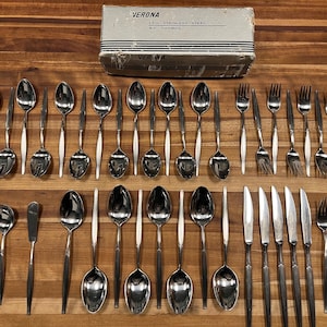 May include: A complete Verona 13-piece stainless steel flatware set by Cosmos, arranged on a wooden surface. The set includes spoons, forks, knives, and a butter knife. The original box is visible in the background, with the text "VERONA 13-pc. STAINLESS STEEL BY COSMOS."