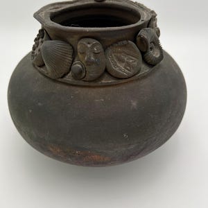 May include: A dark brown ceramic pot with a wide opening and a rounded body. The rim is decorated with seashell and face-shaped embellishments. The pot has a rustic, aged appearance, suggesting it is handmade.