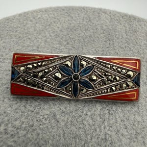 May include: Rectangular silver brooch with a red enamel border and a central design featuring a blue flower and marcasite accents. The brooch has a geometric pattern and is likely vintage or antique.