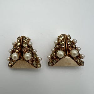 May include: A pair of gold-toned clip-on earrings. Each earring is a triangle shape, adorned with small, sparkling crystals and faux pearls. The earrings have a vintage aesthetic.