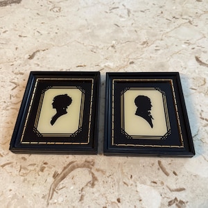 May include: Two framed silhouette portraits. Each black frame has a gold trim and a cream-colored background. One silhouette depicts a woman with an updo, and the other depicts a man with a beard and a high collar. The frames are rectangular.