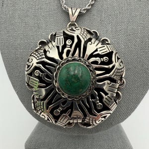 Op de afbeelding: Een zilveren hangerketting met een grote, cirkelvormige hanger. De hanger heeft een ingewikkeld ontwerp met een centrale groene steen en gedetailleerde patronen. De ketting heeft een zilveren ketting.