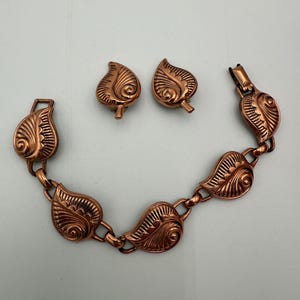 May include: A vintage copper-toned jewelry set featuring a bracelet and matching clip-on earrings. The bracelet and earrings are adorned with leaf-shaped designs, showcasing intricate detailing and a warm, metallic finish. The bracelet measures approximately 7 inches in length.