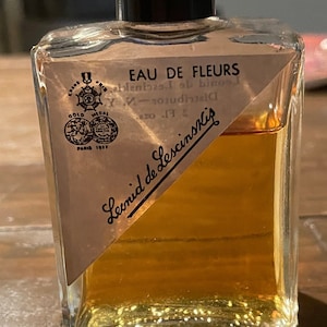 May include: A vintage perfume bottle with a black cap and a clear, rectangular glass body. The bottle contains a golden-colored liquid and features a label with the text "EAU DE FLEURS" and "Leonid de Lescinskis". The label also includes a gold medal emblem.