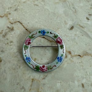 May include: A silver-toned circular brooch with a floral design. The brooch features pink and blue flowers with green leaves, set against a white background. The pin is a classic accessory.