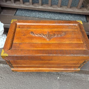 Puede incluir: Un ba&uacute;l rectangular de madera con esquinas de lat&oacute;n. El ba&uacute;l tiene un dise&ntilde;o floral tallado en la tapa y est&aacute; hecho de madera te&ntilde;ida. El ba&uacute;l est&aacute; sobre una superficie gris.