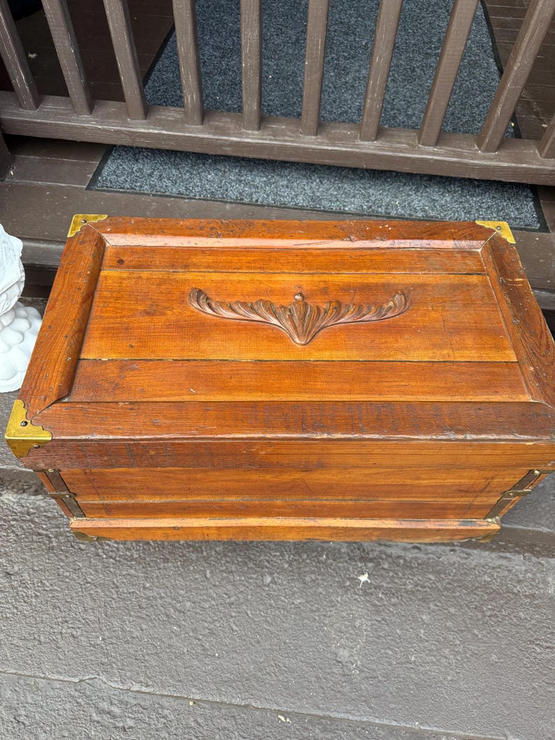 Puede incluir: Un ba&uacute;l rectangular de madera con esquinas de lat&oacute;n. El ba&uacute;l tiene un dise&ntilde;o floral tallado en la tapa y est&aacute; hecho de madera te&ntilde;ida. El ba&uacute;l est&aacute; sobre una superficie gris.