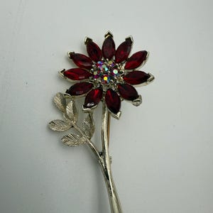 May include: A floral brooch featuring a flower design with deep red, marquise-cut stones as petals. The center of the flower has iridescent stones. The brooch has a silver-tone stem and leaves.