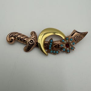 Puede incluir: Broche decorativo con forma de daga con una hoja curva. El broche presenta un mango y una hoja de color cobre, una media luna dorada y detalles turquesa y perlados. Una cuenta roja está en el centro de la flor.