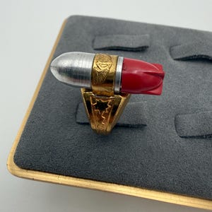 Könnte beinhalten: Ein goldfarbener Ring mit einem horizontalen, patronenförmigen Schmuckelement. Die Patrone hat eine silberfarbene Metallspitze, ein goldenes Mittelband mit geprägtem Adlerdetail und ein rotes Kunststoffheck. Die Ringschiene zeigt Stern- und Zickzackgravuren.