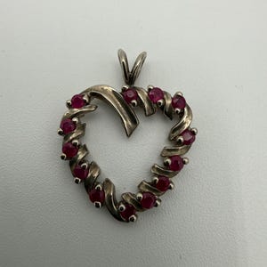 May include: A heart-shaped pendant crafted from a silver-toned metal, featuring a swirling ribbon design. The pendant is accented with ten small, round, deep red gemstones set along the curved frame, complete with a top bail for hanging.
