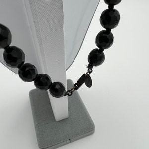 May include: A black faceted bead necklace displayed on a white mannequin. The beads are large and oval-shaped, with a glossy finish. The necklace is fastened with a dark metal clasp. A small tag is attached to the clasp.