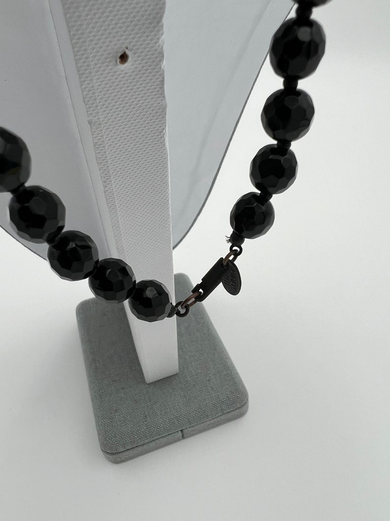 May include: A black faceted bead necklace displayed on a white mannequin. The beads are large and oval-shaped, with a glossy finish. The necklace is fastened with a dark metal clasp. A small tag is attached to the clasp.