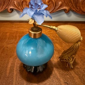 May include: A vintage perfume bottle with a light blue, spherical body and a blue flower-shaped stopper. The bottle has a gold-colored metal collar and a gold-colored atomizer with a tassel. The base is black and the bottle is sitting on a wooden surface.