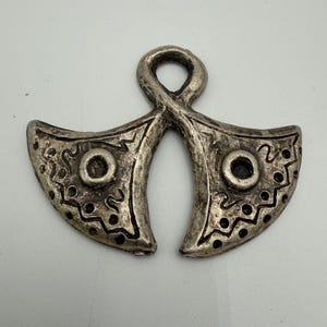 May include: A silver-toned pendant with an abstract design. The pendant features two curved, wing-like shapes connected by a loop at the top. Each wing has a central circular element and decorative patterns. The pendant is likely made of metal.