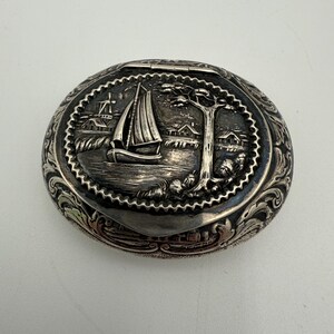 May include: Oval silver-toned trinket box with a detailed nautical scene on the lid. The scene includes a sailboat, a windmill, and a tree. The box has ornate floral and scrollwork around the edges.